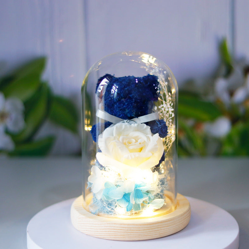 Blue teddy bear and white roses under a glass dome on a wooden base with a blurred floral background.
