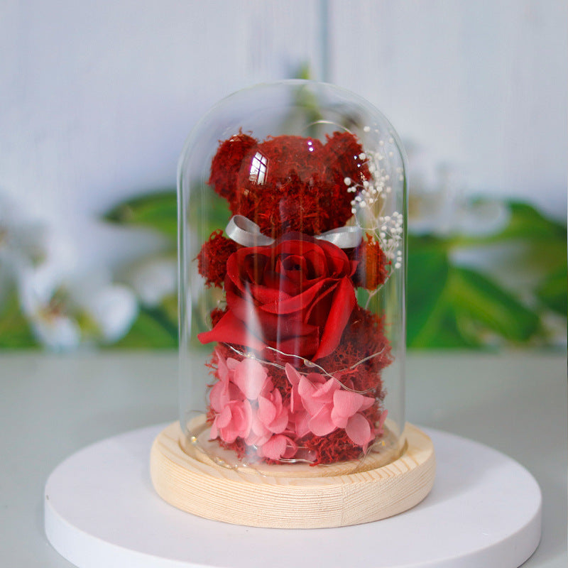 Red teddy bear and flowers inside a glass dome on a white base with a blurred green and white background
