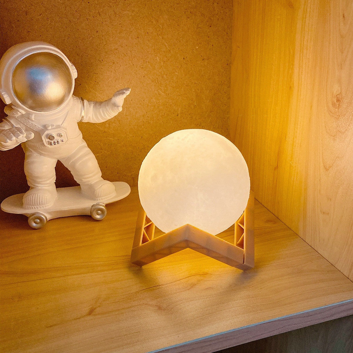 Small astronaut figurine on a skateboard next to a moon lamp on a wooden surface.